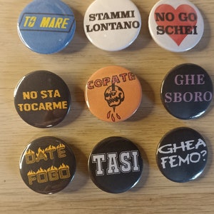 Può includere: Nove bottoni rotondi con diverse frasi e disegni. I bottoni sono neri, bianchi, blu, rossi, arancioni e viola. Le frasi includono "To Mare", "Stami Lontano", "No Go Schei", "No Sta Tocarme", "Copate", "Ghe Sboro", "Date Fogo", "Tasi" e "Ghea Femo?"