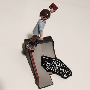 Può includere: Una figurina giocattolo di una persona che indossa una camicia blu e tiene una carta rossa, che esegue un trucco di skateboard su una rampa grigia con una toppa nera e bianca "Vegan Off The Meat".