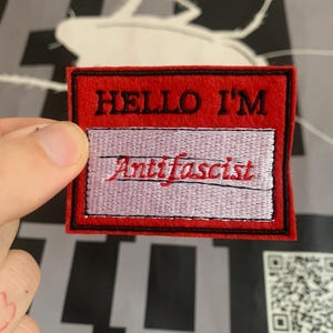 Può includere: Toppa in feltro rosso con cuciture nere. La toppa ha un rettangolo bianco con il testo "HELLO I'M" in nero e il testo "Anti-fascist" in rosso.