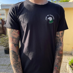 Può includere: T-shirt nero con la scritta ricamata "I poop on fascists" e un disegno di anatra. L'anatra ha la testa bianca, il collo verde e il corpo bianco. La maglietta è realizzata in un materiale morbido.
