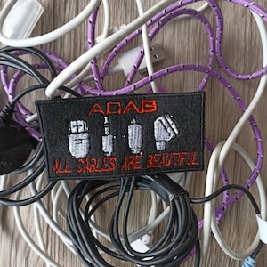 Può includere: Una toppa nera con icone ricamate bianche di diversi tipi di cavi e il testo "ACAB ALL CABLES ARE BEAUTIFUL" in rosso. La toppa è circondata da un groviglio di cavi bianchi, viola e neri.