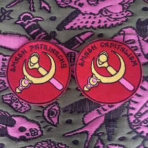 Può includere: Due toppe ricamate rosse con un simbolo di falce e martello giallo. Il testo sulle toppe recita "Smash Patriarchy" e "Smash Capitalism".