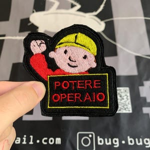 Può includere: Toppa ricamata raffigurante un personaggio dei cartoni animati con un casco giallo e una camicia rossa, con la scritta "POTERE OPERAIO" in rosso su sfondo nero. La toppa ha un bordo nero con cuciture gialle.