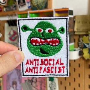 Könnte beinhalten: Gestickter Aufnäher mit einer grünen Cartoon-Figur mit roten Zähnen und Augen. Der Aufnäher trägt den Schriftzug "ANTI SOCIAL ANTI FASCIST" in roter Schrift auf weißem Grund, mit einem schwarzen Rand.