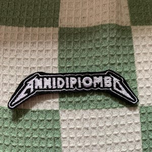 Può includere: Una toppa ricamata nera con scritte bianche che riportano ANNIDIPIOMBE in un carattere angolare stilizzato. La toppa presenta una forma distinta con bordi affilati ed estremità appuntite, con un dettagliato lavoro di filo bianco su fondo nero.