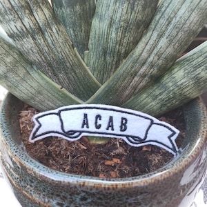 May include: White embroidered patch with black stitching in the shape of a banner with the text "ACAB" on a green plant in a brown pot.