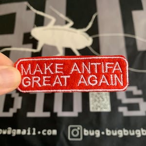 Può includere: Una toppa rossa con testo ricamato bianco che recita "MAKE ANTIFA GREAT AGAIN". La toppa è rettangolare con un bordo bianco. Lo sfondo è uno striscione nero con grafica e testo bianchi.