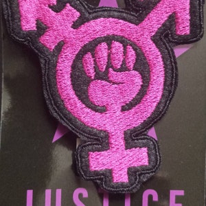 Può includere: Una toppa ricamata rosa e nera con un pugno chiuso all'interno di un cerchio, un simbolo transgender e la parola "JUSTICE" sotto.