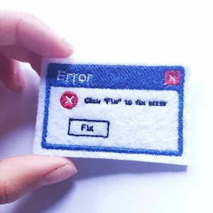 Può includere: Una toppa di feltro bianco con un bordo blu e la parola "Error" in alto. La toppa presenta un'icona "X" rossa e il testo "Click 'Fix' to fix error". Un pulsante bordato di nero recita "Fix".