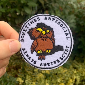 Può includere: Una toppa ricamata bianca con un gufo marrone con occhi gialli. Il testo intorno al gufo dice "Sometimes Antisocial Always Anti-Fascist".