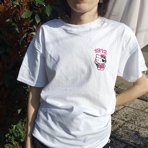 Può includere: Maglietta bianca con un'immagine rosa di Hello Kitty che tiene un telefono e il testo "1312" sopra di lei.