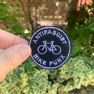Può includere: Una toppa circolare nera con testo ricamato bianco e una grafica di bicicletta. Il testo recita "ANTIFASCIST BIKE PUNX" in un motivo circolare attorno all'illustrazione della bicicletta.