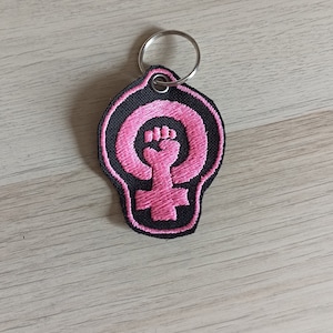 Può includere: Portachiavi con un simbolo femminista rosa con un pugno alzato. Il simbolo è ricamato su uno sfondo in tessuto nero.