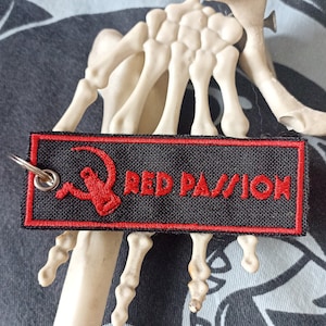 Può includere: Un portachiavi in tessuto nero con cuciture rosse e il testo "RED PASSION" in rosso. Un simbolo di falce e martello stilizzato rosso si trova a sinistra del testo.