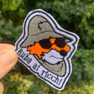 Può includere: Una toppa ricamata bianca con un personaggio dei cartoni animati che indossa un cappello grigio e occhiali da sole. Il personaggio ha i capelli arancioni e un viso bianco. Il testo "Ruba at Ricci" è ricamato in nero sulla toppa.