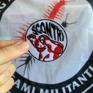 Può includere: Una toppa rotonda ricamata bianca con la scritta "SCONTRI" in nero, testo curvo. La toppa presenta un personaggio dei cartoni animati rosso e bianco. Sullo sfondo una bandiera con testo nero e un disegno di millepiedi rosso.
