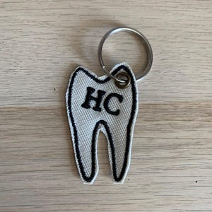 Può includere: Un portachiavi in tessuto a forma di dente, con le lettere "HC" ricamate in nero. Il dente è bianco con un contorno nero ed è attaccato a un portachiavi argentato.