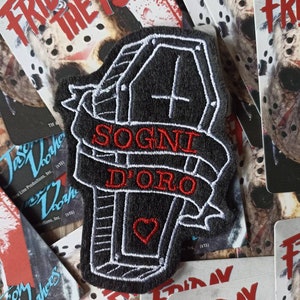 Può includere: Un ricamo nero e bianco con una bara, un nastro rosso con la scritta "SOGNI D'ORO" e un cuore rosso. Il ricamo è circondato da diverse carte promozionali del film "Venerdì 13".