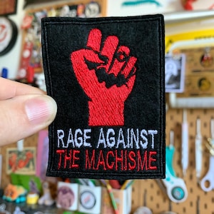 Può includere: Toppa quadrata nera con un pugno rosso e le parole "RAGE AGAINST THE MACHISME" ricamate in bianco e rosso. La toppa è tenuta contro uno sfondo disordinato.