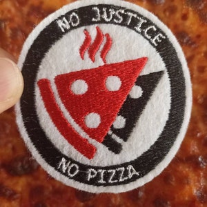 Può includere: Una toppa rotonda bianca con la scritta "NO JUSTICE NO PIZZA" in nero. Il design presenta una fetta di pizza rossa con fori bianchi e una fetta di pizza nera. Sopra, tre fiamme rosse.