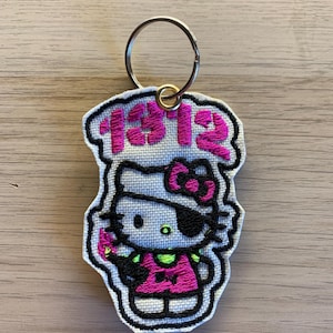 Può includere: Portachiavi ricamato con un gatto dei cartoni animati con una benda sull'occhio e un vestito rosa. Il portachiavi ha un anello argentato e il numero "1312" in rosa. Il gatto è bianco con un occhio nero e un fiocco rosa.