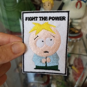 Può includere: Una toppa ricamata bianca con bordo nero che presenta un personaggio dei cartoni animati con capelli biondi, una camicia blu e pantaloni verdi. Il testo "FIGHT THE POWER" è sopra il personaggio.
