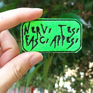 Può includere: Un cerotto di tessuto verde con testo ricamato nero che recita "NERVY" e "BAD" "ASS".