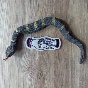 Può includere: Un giocattolo a forma di serpente nero e giallo con una toppa ricamata bianca che raffigura una mano che tiene un serpente con una corona in testa. La toppa ha accenti rossi e verdi.