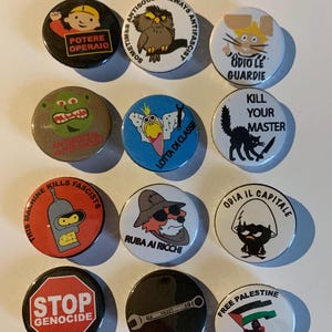Può includere: Una collezione di spille rotonde con vari disegni e testi. Le spille presentano personaggi dei cartoni animati, slogan politici e illustrazioni. I colori includono rosso, nero, bianco e marrone. Alcune spille hanno testo in italiano.