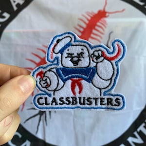 Può includere: Toppa ricamata con un personaggio dei cartoni animati in tenuta da marinaio, con la scritta "CLASSBUSTERS". Il personaggio è bianco con dettagli blu e rossi. Lo sfondo include testo e un'illustrazione rossa.