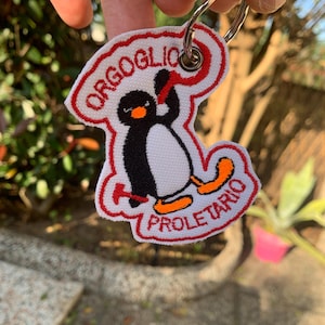 Könnte beinhalten: Ein weißer Stoffschlüsselanhänger mit rotem, gesticktem Rand. Der Schlüsselanhänger zeigt einen Comic-Pinguin, der einen roten Schal trägt und einen roten Hammer hält. Der Text "ORGOGLIO PROLETARIO" ist in Rot auf den Schlüsselanhänger gestickt.