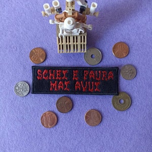 Può includere: Una scultura Lego con una base a forma di gabbia e multiple sporgenze bianche e marroni. Una toppa nera con testo ricamato in rosso, "SOHEI E FAURA MAI AVUI", è circondata da varie monete, tra cui centesimi e dimes, su una superficie viola.