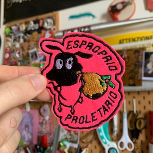 Può includere: Una toppa ricamata rosa con un cartone animato di una pecora che trasporta un frutto dorato. La toppa presenta le parole "ESPROPRIO" e "PROLETARIO" in rosso. La pecora ha la faccia nera ed è bordata di nero.
