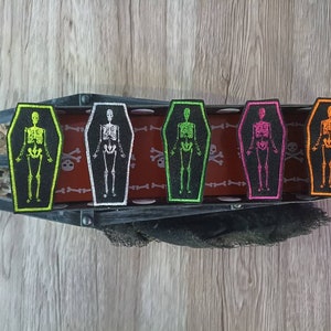 Può includere: Cinque toppe ricamate da stirare a forma di bara con scheletri all'interno. Le bare sono nere con un contorno bianco. Gli scheletri sono di vari colori: verde, bianco, rosa, arancione e rosso.