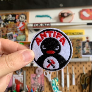 Può includere: Una toppa rotonda, bianca e nera con un disegno di pinguino e la scritta "ANTIFA" in rosso. Il pinguino ha un becco rosso e un simbolo di martello e chiave inglese incrociati. La toppa è tenuta da una mano.