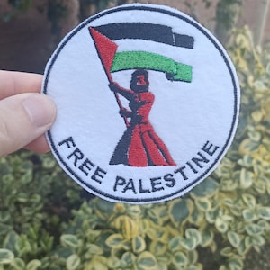 Può includere: Una toppa circolare bianca con ricamo nero. La toppa presenta una donna che tiene una bandiera palestinese con le parole "FREE PALESTINE" ricamate sotto.