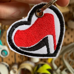 Può includere: Un portachiavi a forma di cuore rosso e nero con un design di scarpa con tacco alto rosso. Il portachiavi ha un anello di metallo argentato.