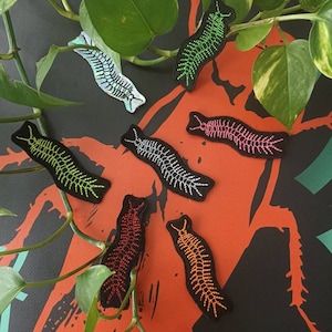 Può includere: Un set di sette toppe ricamate da stirare con un design a millepiedi in vari colori, tra cui verde, rosa, argento, nero, arancione e rosso. Le toppe sono esposte su uno sfondo nero e rosso.