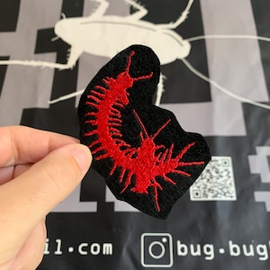 Op de afbeelding: Zwarte geborduurde patch met een rood duizendpootontwerp. De patch wordt omhoog gehouden tegen een zwarte achtergrond met witte tekst en een kakkerlakafbeelding. De Instagram-handle "bug.bug" is zichtbaar.