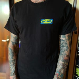 Può includere: Una t-shirt nera a maniche corte con una toppa rettangolare sul petto sinistro. La toppa presenta uno sfondo blu con un ovale giallo al centro contenente il testo blu ACAB in un carattere grassetto senza grazie, imitando un logo.