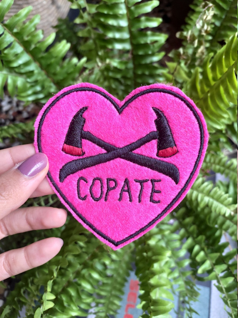 Puede incluir: Un parche de fieltro rosa en forma de coraz&oacute;n con hachas cruzadas bordadas en negro y la palabra "COPATE" en letras negras.