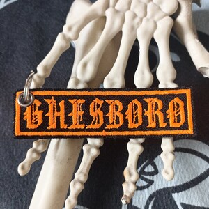 Può includere: Portachiavi ricamato nero e arancione con la parola "GHEISBORO" in caratteri gotici.