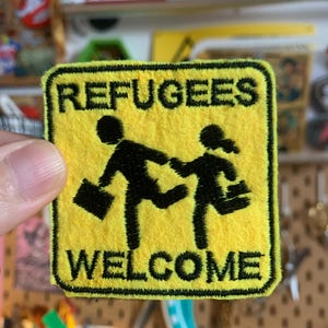 Può includere: Toppa in tessuto giallo con testo e immagini ricamate in nero. Il testo recita "Refugees Welcome" e raffigura due figure stilizzate che camminano con valigie.