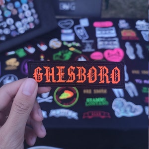 Può includere: Una toppa rettangolare nera con la scritta "GHESBORO" in arancione, con lettere in stile gotico. La toppa è tenuta davanti a un tavolo coperto da varie toppe e spille. Lo sfondo è sfocato, suggerendo un'ambientazione di mercato all'aperto.