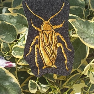 Può includere: Un cerotto di feltro nero con un design di scarafaggio dorato ricamato. Lo scarafaggio è dettagliato con gambe e corpo segmentati.
