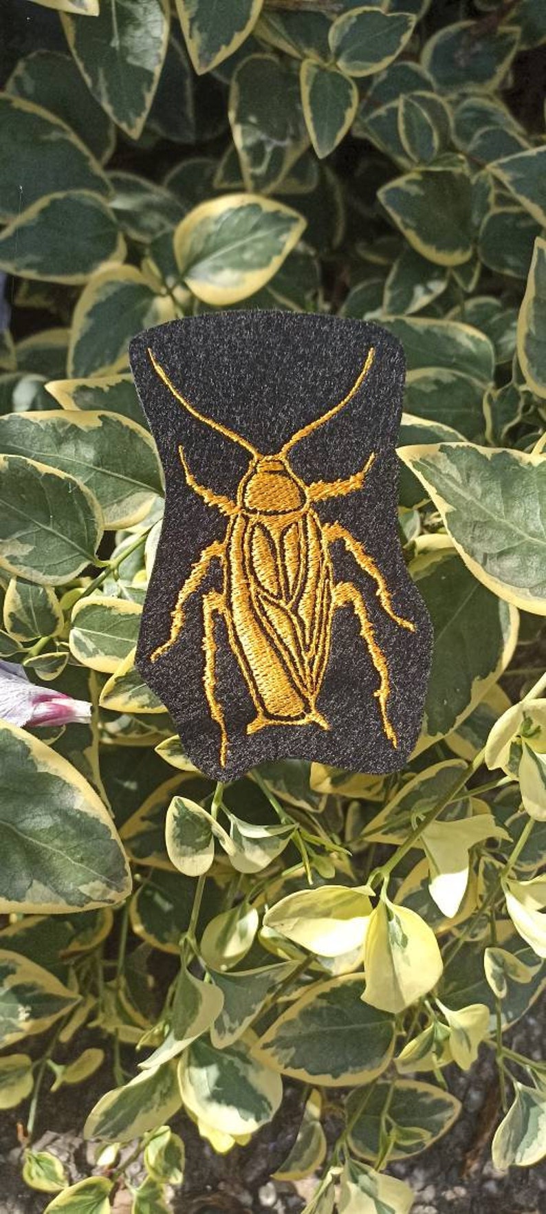 Op de afbeelding: Een zwarte viltpatch met een geborduurd gouden kakkerlakontwerp. De kakkerlak is gedetailleerd met gesegmenteerde poten en een lichaam.