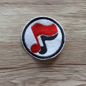 Può includere: Una toppa rotonda bianca con un bordo nero. La toppa presenta una nota musicale rossa e nera con una bandiera rossa in cima.