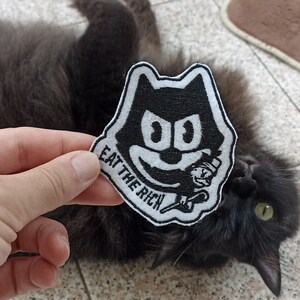 Può includere: Toppa ricamata in bianco e nero con un gatto dei cartoni animati che tiene una tazza di caffè con il testo "Eat the Rich" sotto.