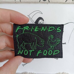 Può includere: Toppa ricamata nera con cuciture verdi. La toppa presenta un maiale, una gallina e il testo "FRIENDS NOT FOOD".