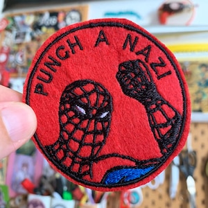 Può includere: Una toppa circolare ricamata di colore rosso con una grafica in filo nero raffigurante una maschera da supereroe e un pugno alzato. Il testo PUNCH A NAZI è ricamato in nero lungo la curva superiore della toppa, che presenta anche accenti blu.